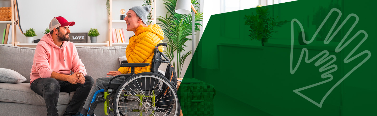 Header: Eine Betreuungsperson sitzt auf einem Sofa und unterhält sich mit einer Person im Rollstuhl